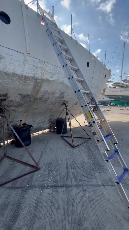 Long aluminum ladder leaning against Tortuga in the boatyard, with the hull towering above the ground.