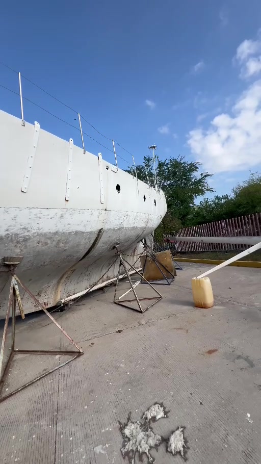 Long view along Tortuga’s white hull, with round portlights, supporting stands and blue sky.
